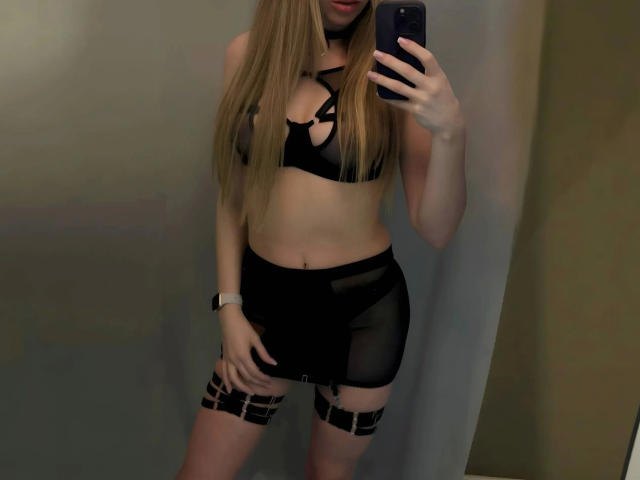 EngelEva-hot with long blonde hair, wearing black strappy bra, sheer skirt, and black garter, taking a mirror selfie with phone, showing her bare tits and ass.