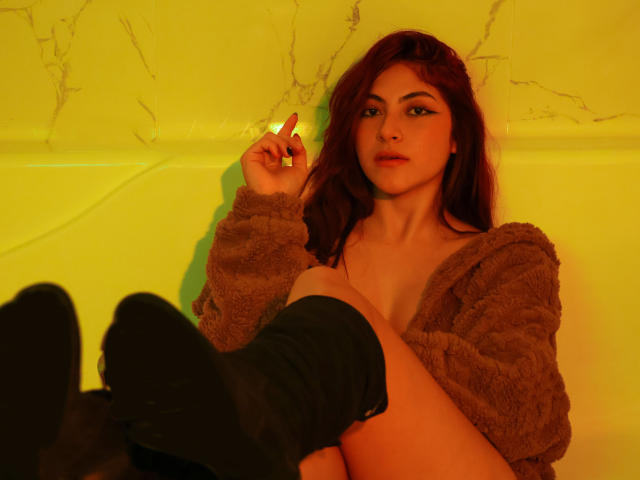 SpencerLe, com cabelo ruivo, olhos verdes e lábios carnudos, vestindo uma jaqueta marrom felpuda, meias pretas e sem calças, segurando um cigarro, iluminada em amarelo, sentada com as pernas levantadas..