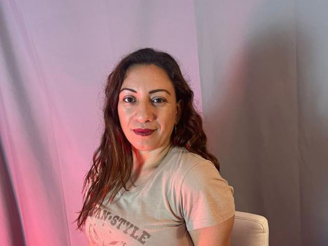 Britney69 with wavy brown hair, wearing a gray T-shirt, has dark red lipstick and a slight smile, seated against a pink and gray curtain.