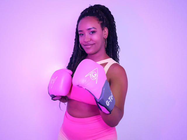 NaomiReyes, dark-skinned woman with long braids, wearing pink boxing gloves, pink and yellow sports bra, pink high-waisted pants, smiling, holding gloves in front, purple-pink lighting.