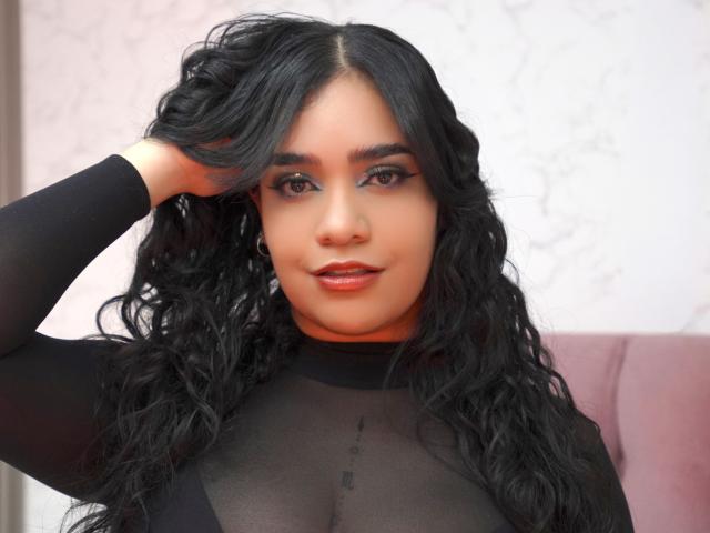 Alicia, mit lockigem, schwarzem Haar, trägt ein durchsichtiges schwarzes Oberteil, ihre Hand ist in ihrem Haar, sie hat braune Augen und glänzende, rote Lippen, und man kann ihre Brüste durch den Stoff erkennen..