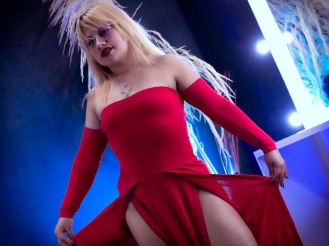 LilithCastelli wearing a red off-shoulder dress with a high slit, holding the skirt, blonde hair, purple glasses, dark lipstick, feathered headpiece, and a small chest tattoo.