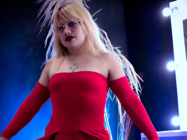 LilithCastelli has blonde hair, red off-shoulder dress, and white feather headpiece, with dark red lipstick, wearing glasses, showing small tits, standing in front of a lit mirror.