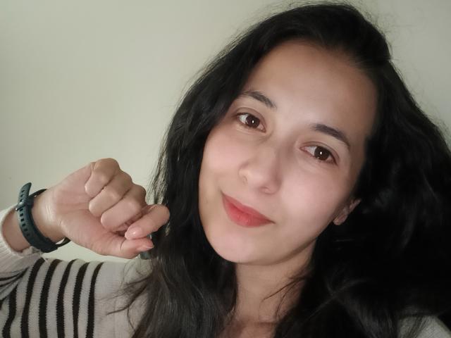 AnneStone with dark wavy hair, brown eyes, and light skin, wearing a striped shirt, holding her finger to her mouth, smiling subtly, wearing a green wristband.
