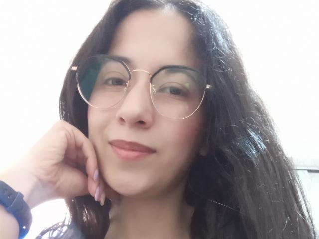 AnneStone with dark, wavy hair, wearing round glasses, slight smile, pink lipstick, resting cheek on hand, blue wristband, light skin, soft facial features, subtle makeup.