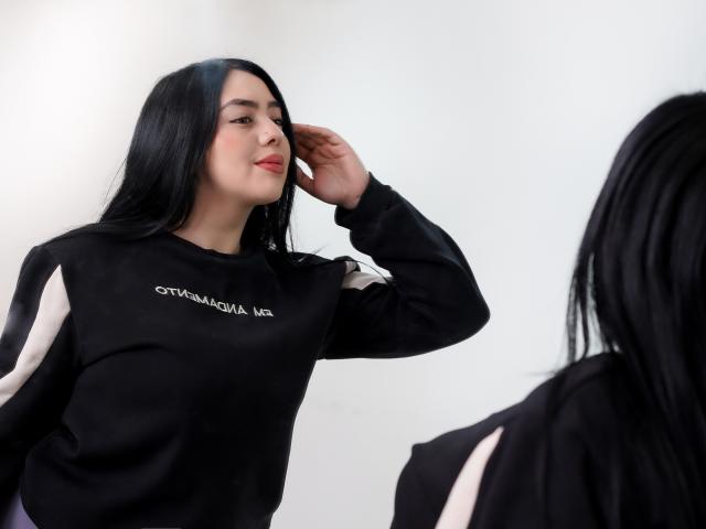 ZaahraLewis with long black hair, wearing a black sweatshirt with white stripes and text, touching her ear, smiling, facing another person with similar black hair in the foreground.