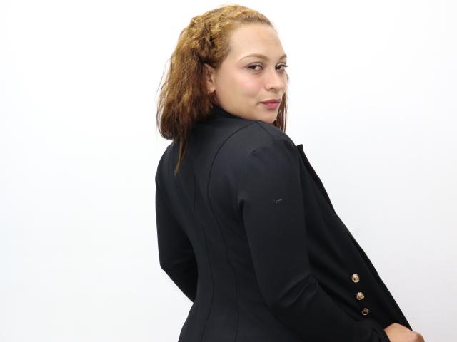 Rhoxana with braided brown hair, black jacket, and gold buttons, looking over her shoulder, showing a subtle smile and her ass. She has light skin and pink lips.
