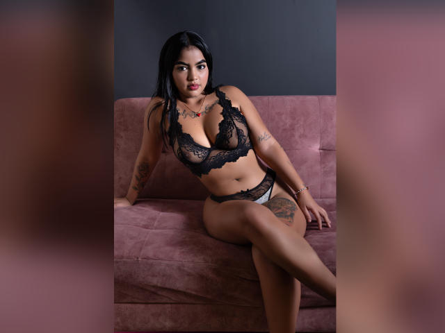 GiaAttum portant un soutien-gorge et une culotte en dentelle noire, assise sur un canapé rose, les jambes croisées, avec des tatouages sur le bras et la cuisse, des cheveux foncés, regardant la caméra, peau mate, collier rouge..