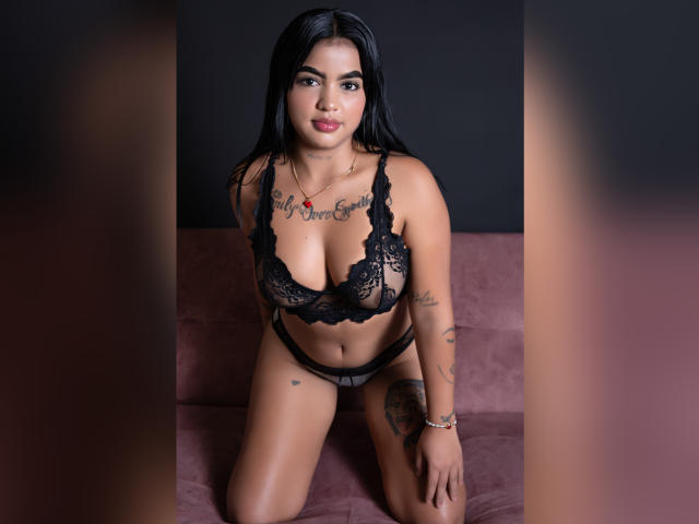 GiaAttum met een zwarte kanten bh en slip, tatoeages en lang zwart haar, knielend op een roze bank, waarbij haar borsten en vulva zichtbaar zijn, kijkend naar de camera met een lichte glimlach..