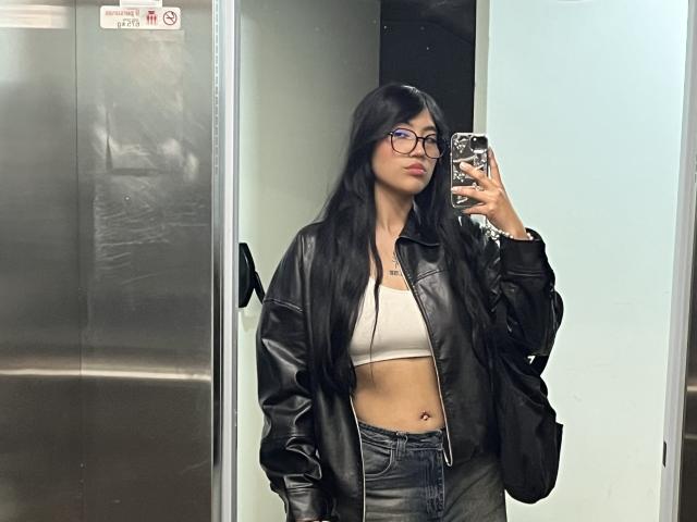 Elizaa with long black hair, glasses, and a leather jacket, showing her tits in a beige bra and denim pants, taking a mirror selfie in a metal elevator.