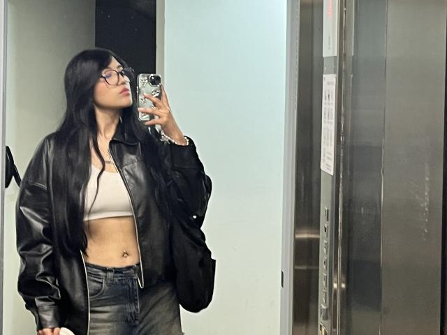 Elizaa with long black hair, glasses, a leather jacket, and a beige bralette, showing her flat stomach and belly button, taking a mirror selfie in a elevator.