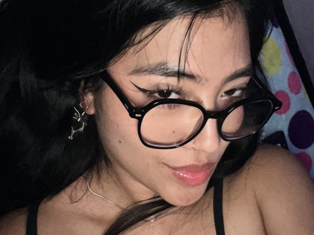 Elizaa with black glasses, dark hair, and a nose piercing, wearing a black top, showing a small mole on her cheek and a slight smirk. Boobs partially visible.