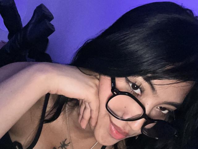Elizaa with black hair, wearing glasses, has her hand on her chin, showing a tattoo on her chest, and a subtle smile, in a dimly lit, purple-hued setting.