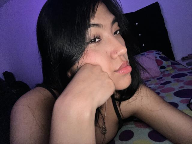 Elizaa with black hair, nose ring, and long eyelashes, resting her head on her hand, showing her pink lips and smooth skin, wearing a black top, on a bed.