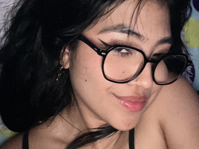 Elizaa with black glasses, dark wavy hair, and a cute smile, showing a small beauty mark near her nose. She's wearing a black strap and has a subtle nose ring.