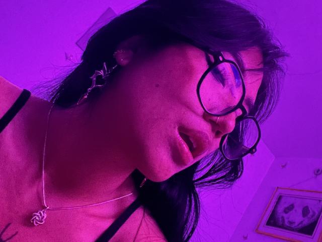 Elizaa with purple-tinged skin, black glasses, and black hair, wearing a black top, slightly open mouth, and a silver necklace with a cross pendant, looking down.