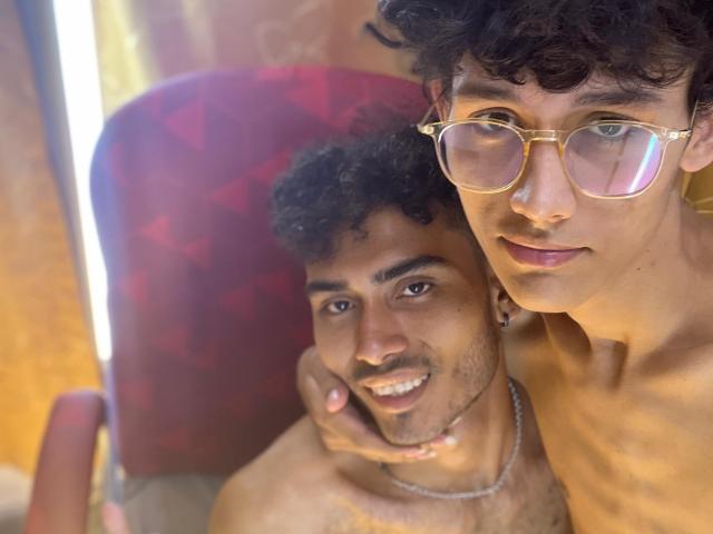 Two shirtless Alenders with curly black hair, one wearing glasses, are in a red chair. The smiling Alender has a necklace, the other looks down. Sunlight is on their faces.
