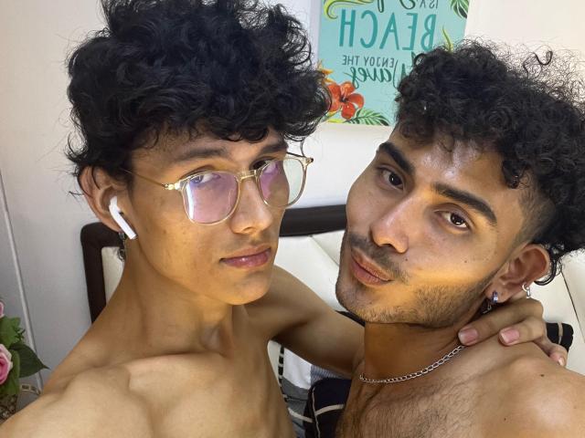 Two shirtless Alenders with curly black hair, wearing glasses and earbuds, looking at the camera. One has a necklace, the other a slight beard. Their faces are close together.