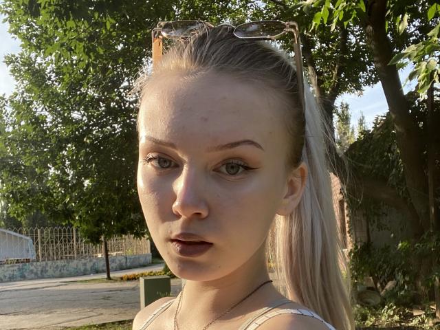 KaroasLove has light skin, green eyes, and long blonde hair in a ponytail. She's wearing sunglasses on her head and a white tank top, with a serious expression.
