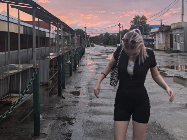 KaroasLove with blonde ponytail, black short-sleeve shirt, and black shorts, walking on wet, reflective sidewalk, looking down, with striped bag over shoulder, at sunset.