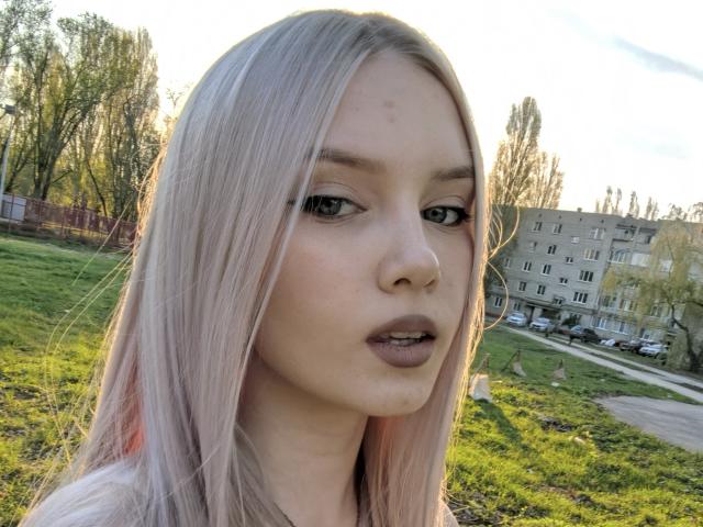 KarosLove has pale pink hair, green eyes, and a slightly open mouth with brown lipstick. Her face shows a small zit. She's looking at the camera.