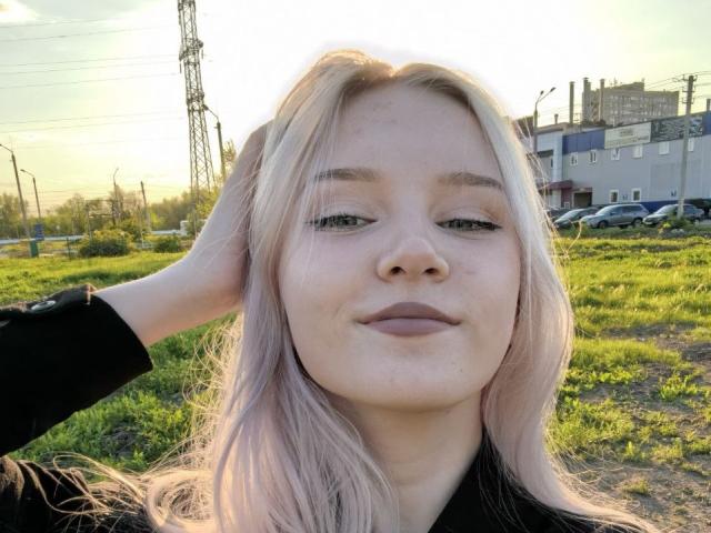 KaroasLove has pale pink hair, green eyes, and light skin. She's wearing a black top, hand in hair, smiling slightly, and has a small nose with subtle makeup.