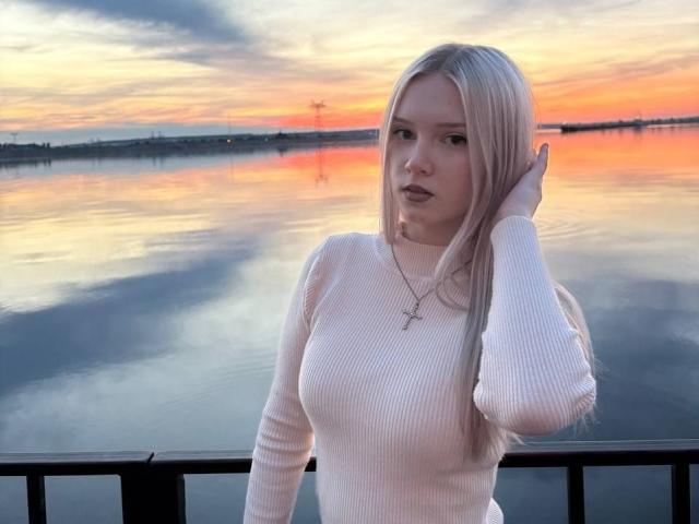 KaroasLove with long blonde hair, wearing a white ribbed sweater, has a cross necklace and a serious look, standing by a black railing at sunset with reflective water.