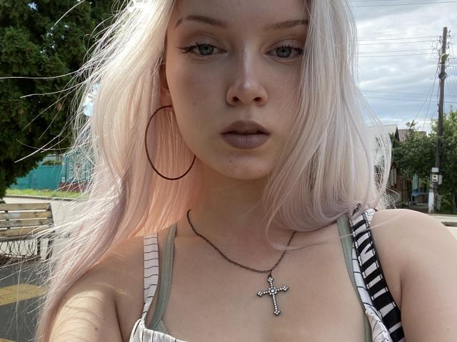 KarosLove with pale pink hair, wearing large hoop earrings, a cross necklace, and striped tank top, has pouty lips and a serious expression. Her tits are small and subtle.