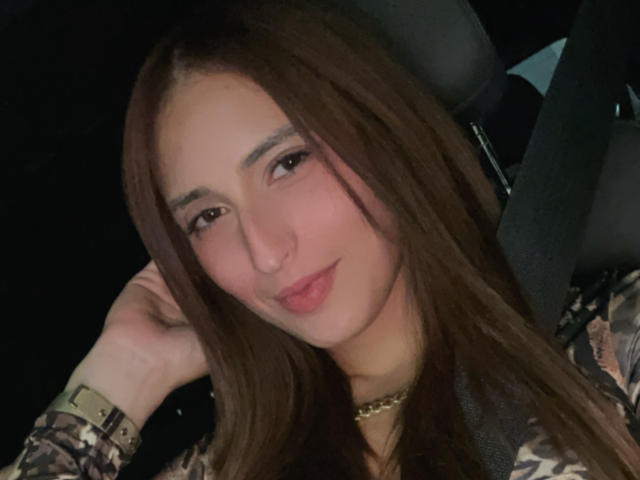 TastyBabex has long brown hair, light skin, and is smiling slightly with her hand near her head. She's wearing a camouflage jacket and a gold chain.
