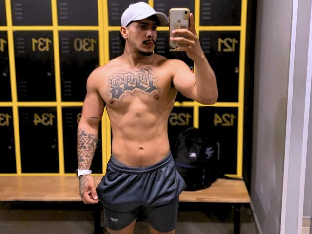 DeimonOfficial, zonder shirt, gespierd, getatoeëerde borst, "DEMON" tatoeage, zwarte korte broek, witte pet, maakt een spiegelzelfportret, kleedkamer, tatoeage op de rechterarm, horloge om de linkerpols, telefoon in de rechterhand..