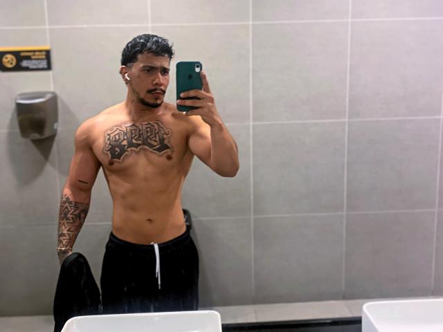 DeimonOfficial, sin camisa, con el pecho y el brazo tatuados, sosteniendo un teléfono en un selfie frente a un espejo en el baño, vistiendo pantalones negros, mostrando un físico musculoso, bronceado y atractivo..