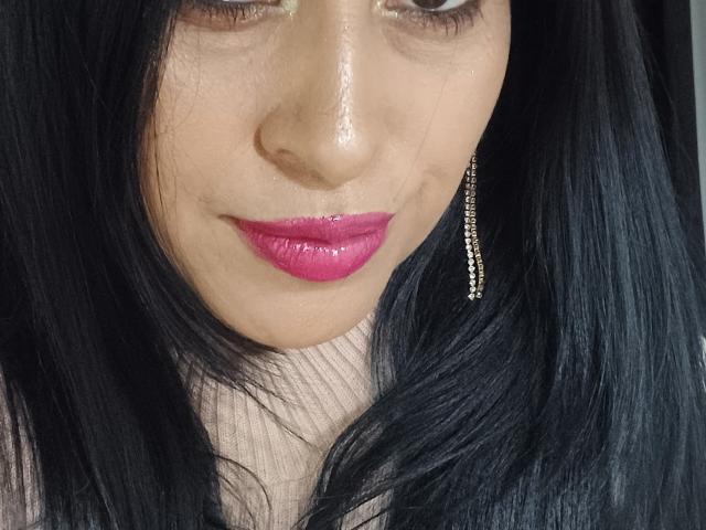 Apasion's face with glossy pink lips, black hair, and a single gold dangling earring. Her nose is slightly shiny, and she's looking down with a subtle smile.