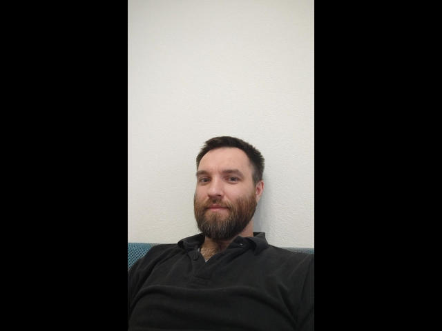 SunColifild, a bearded dude with short dark hair, wearing a black polo, sitting against a white wall, looking slightly to the left with a relaxed, confident expression.