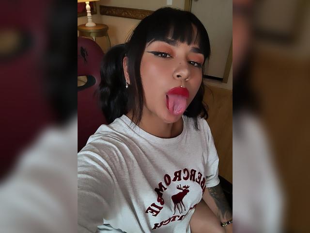 Karina Blake mit schwarzen Zöpfen, die Zunge herausstreckt, trägt ein weißes Hemd mit einem roten Elch darauf. Sie hat auffälligen Eyeliner, roten Lippenstift und einen Nasenring und blickt direkt in die Kamera..