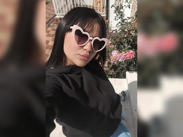 KarinaBlake with heart-shaped pink sunglasses, black hoodie, and blue pants, pouting with dark hair and light skin, standing outdoors with pink flowers in the background.