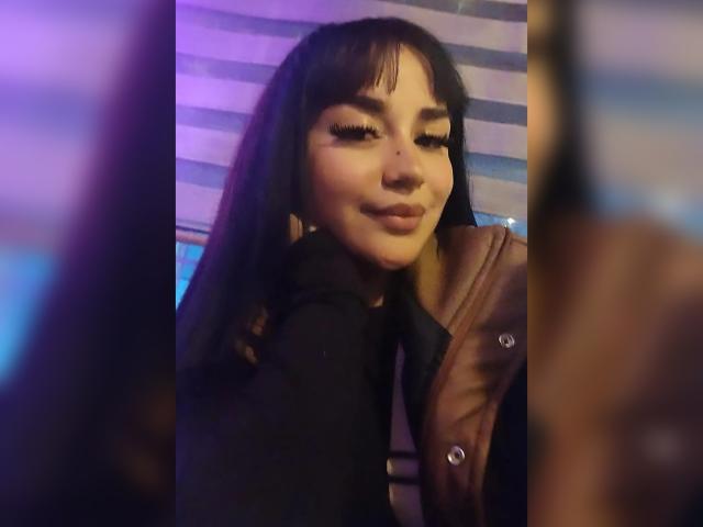 KarinaBlake with long dark hair, heavy eyelashes, and a cute smile, wearing a black top and brown leather jacket, has her ass slightly turned to the camera.