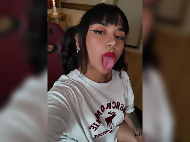 KarinaBlake, with black pigtails and bangs, sticking out her tongue, wearing a white shirt with a red deer, has red lipstick and bold eyeliner, and a tattoo on her arm.