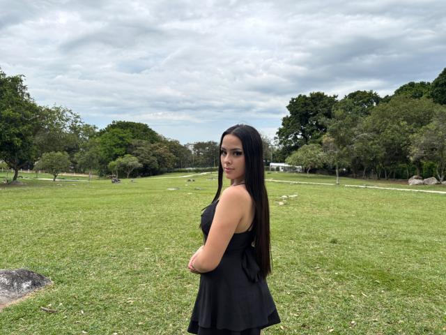 AlisonTatcher stands in a grassy park, facing sideways, showing her black sleeveless top, long dark hair, and a subtle smile, with her arms crossed, under a cloudy sky.
