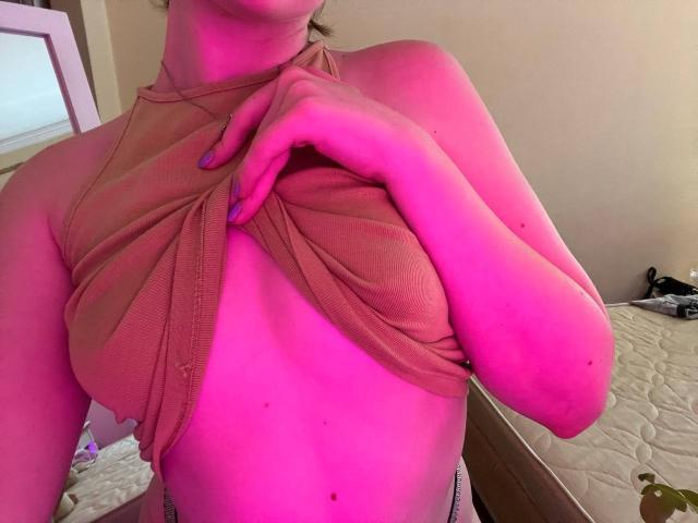 Los senos rosados de Valissia quedan expuestos mientras levanta su camisa marrón con textura vertical con una mano, mostrando su abdomen plano, con pecas y sus uñas pintadas de morado..