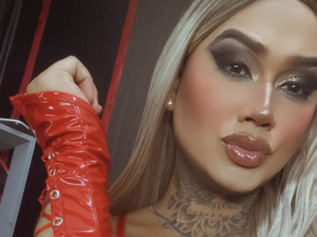 DannikaSaenz with glossy pink lips, dark smoky eye makeup, red vinyl arm, blonde hair, and intricate tattoo on her neck. Boobs not visible, focus on face and arm.