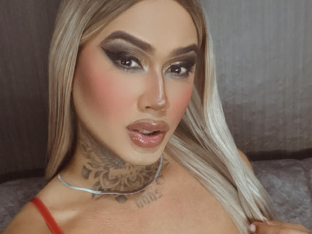 DannikaSaenz has blonde hair, heavy eyeliner, pink blush, glossy lips, and a detailed tattoo on her neck. She's wearing a red top, looking sexy.