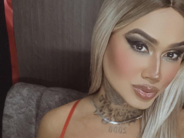 DannikaSaenz with blonde hair, heavy eyeliner, and glossy lips, showing a tattooed neck, wearing a red strap, looking at the camera with a slight pout.