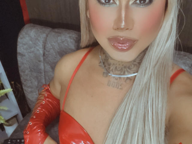 DannikaSaenz with long blonde hair, red latex top, and matching red gloves, glossy pink lips, tattooed neck, and visible cleavage, looking seductively at the camera.