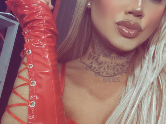 DannikaSaenz with glossy lips, long blonde hair, red leather glove, lace-up front, intricate tattoo on her neck, shiny skin, and a subtle pearl earring. Boobs partially visible.