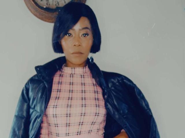 BuffyX con i capelli neri corti, indossa un abito a quadri rosa e una giacca nera, con un'espressione seria, un orologio sullo sfondo, illuminazione soffusa, inquadratura media..