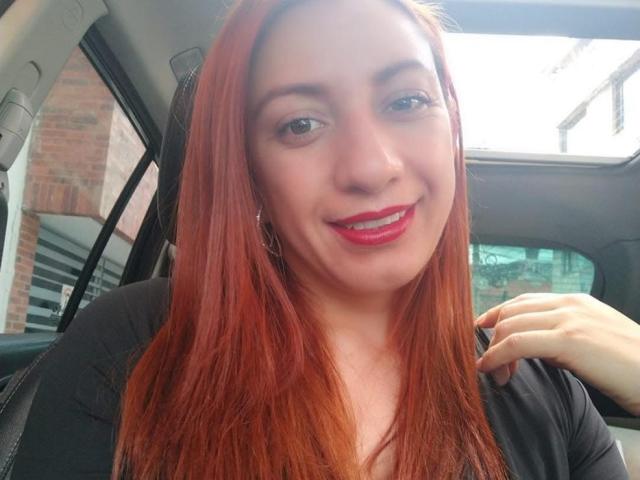 Britney69 with red hair, fair skin, and red lipstick, smiling in a car, wearing a black top, hand near collar, showing her tits subtly, with a sexy look.