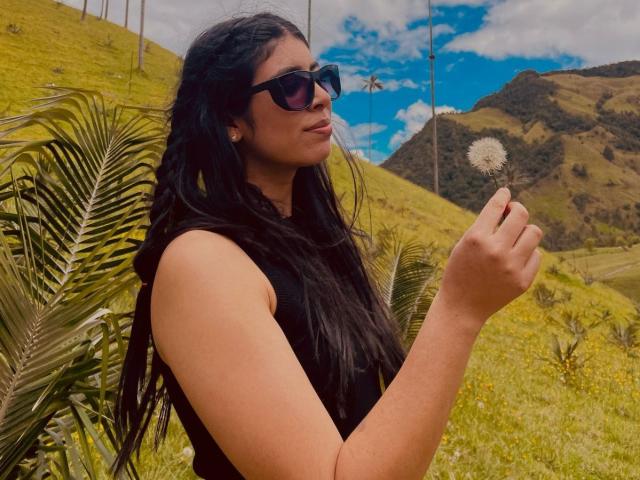 MiluSky with long black hair, wearing black sunglasses and sleeveless black top, holding a dandelion seed, standing in front of green palm leaves and a blue sky.