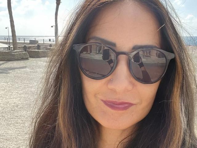 ChiaraNuri with brown hair, wearing black cat-eye sunglasses, has a nose piercing and pink lipstick, reflecting a building in her sunglasses. Her ass is not visible.