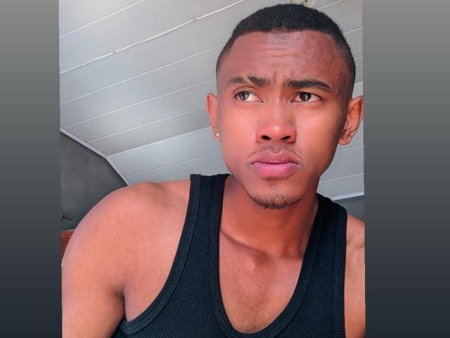 A photo of SugarBoyd with short black hair, a small earring, and a black tank top, making a slightly confused or annoyed facial expression. He's got a medium brown skin tone.