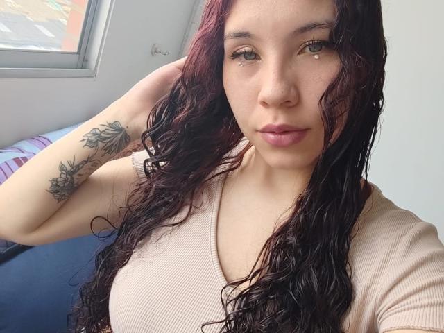 SofiBrandford has wavy dark red hair, brown eyes, a nose piercing, and a tattoo on her left arm. She's wearing a pink ribbed top, showing her tits.